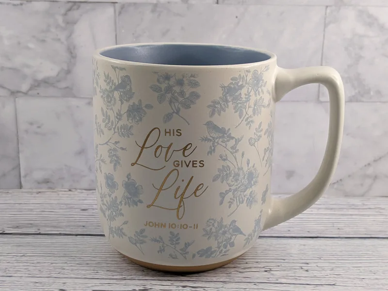 A religious mug that's a cream color with a light blue flower design and a gold Bible verse with a blue interior.
