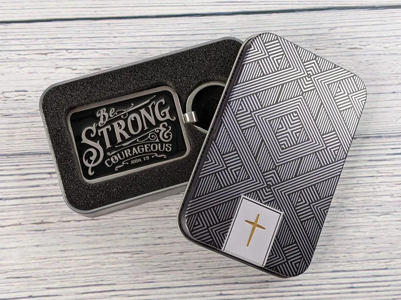 A silver metal Bible verse keyring with a black enamel design and the verse Joshua 1:9, that comes in a gift tin.