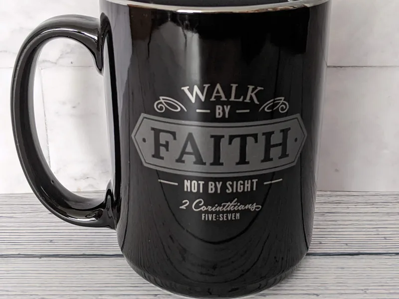 A glossy black mug with a Bible verse: Walk by faith not by sight.