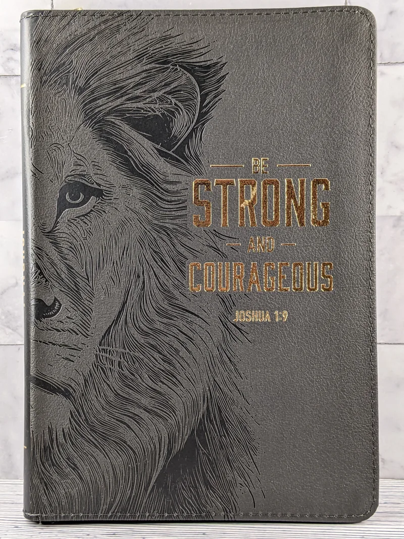 A gray faux leather journal with an image of a lion and a Joshua 1:9 Bible verse on the cover.
