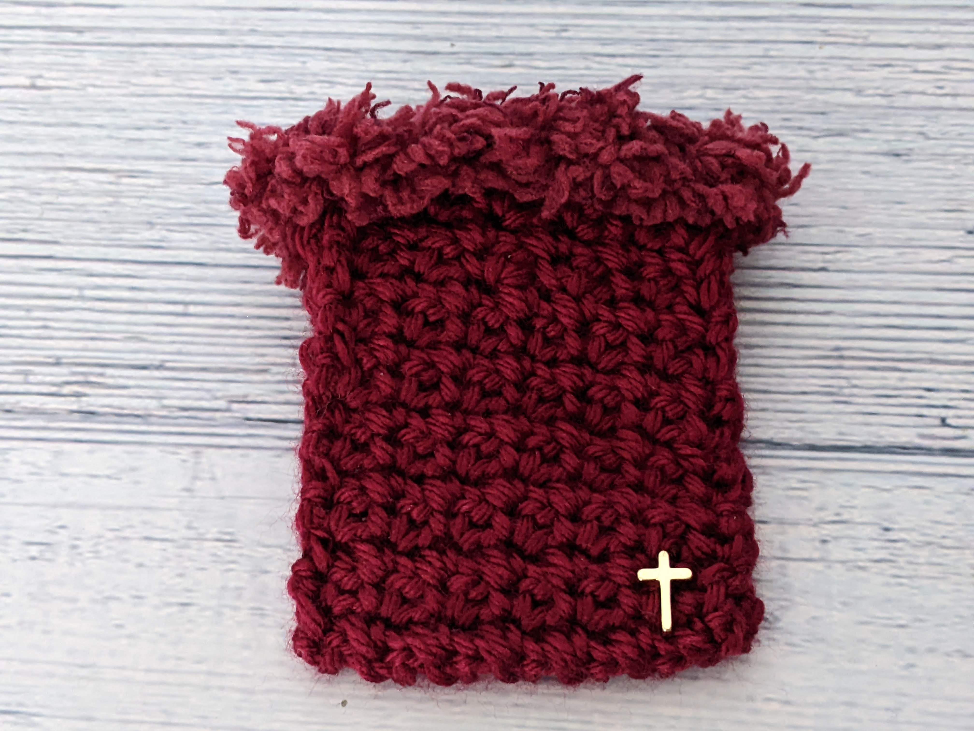 A crocheted burgundy gift card holder bag with fuzzy trim and a golden cross motif.