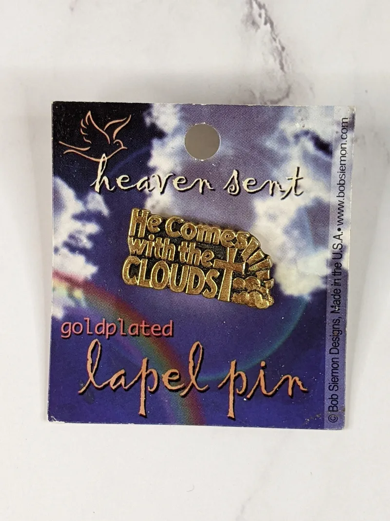 A gold metal lapel pin with a cross design and the words, "He comes with the clouds."