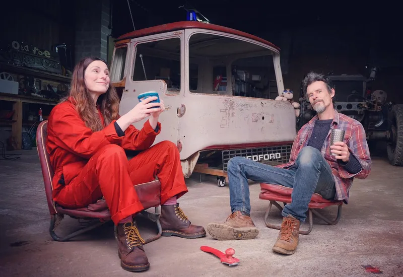 Man and woman having a tea break in a workshop in front of the cab of an old Bedford truck
