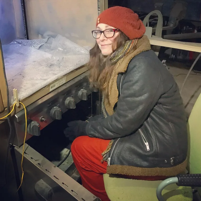 Woman in a coat sitting next to an oven to keep warm