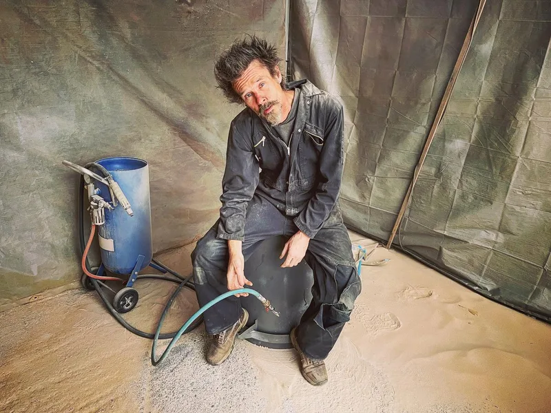 Man in overalls sitting on an upside down bin covered in sand with a sandblaster in his hand