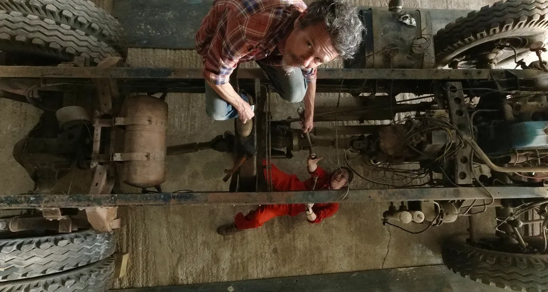 Aerial view of a man standing on a truck chassis frame passing a spanner to a woman in orange overalls lying underneath the truck