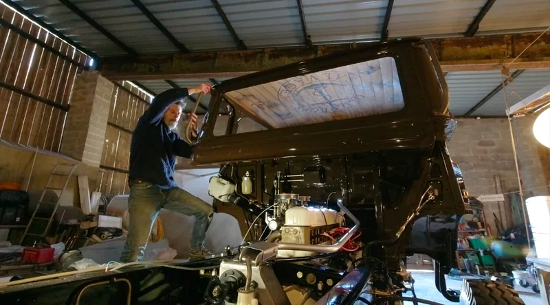Man standing on the back of a brown classic Bedford truck, fitting a grab handle to the cab