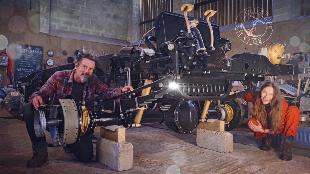 Man and woman with the restored chassis of a 1970s Bedford truck painted in black and gold