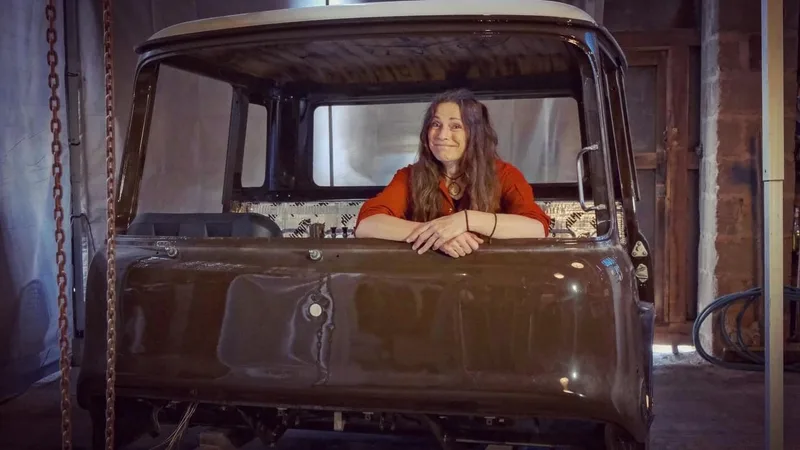 Woman in orange overalls sits inside a brown Bedford truck cab which is being restored