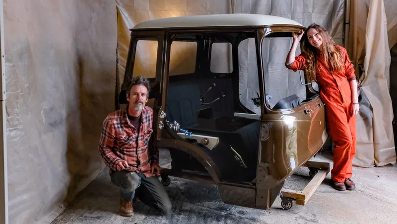 Man and woman in overalls stand with the restored shell of a Bedford truck cab which is painted brown and cream