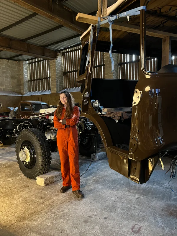 Woman in orange overalls standing next to the separated cab and chassis of a restored classic Bedford truck