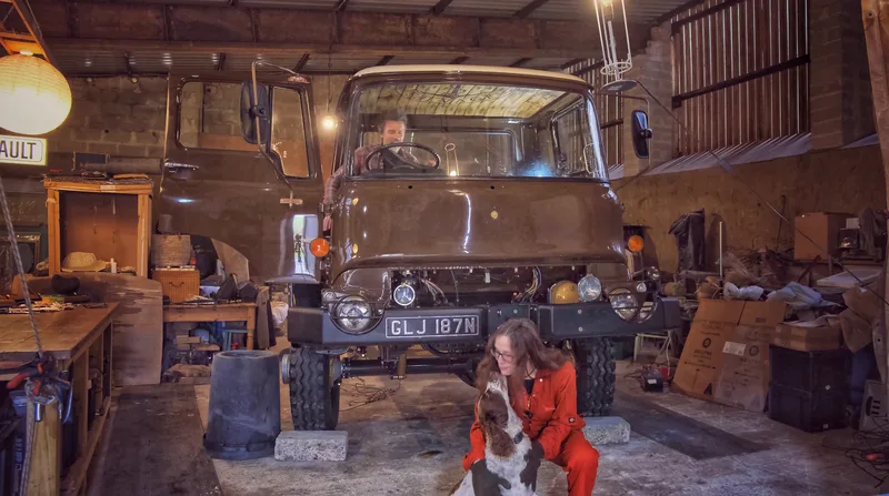Brown Bedford truck in a workshop with a man sitting in the driving seat and a woman playing with a dog 