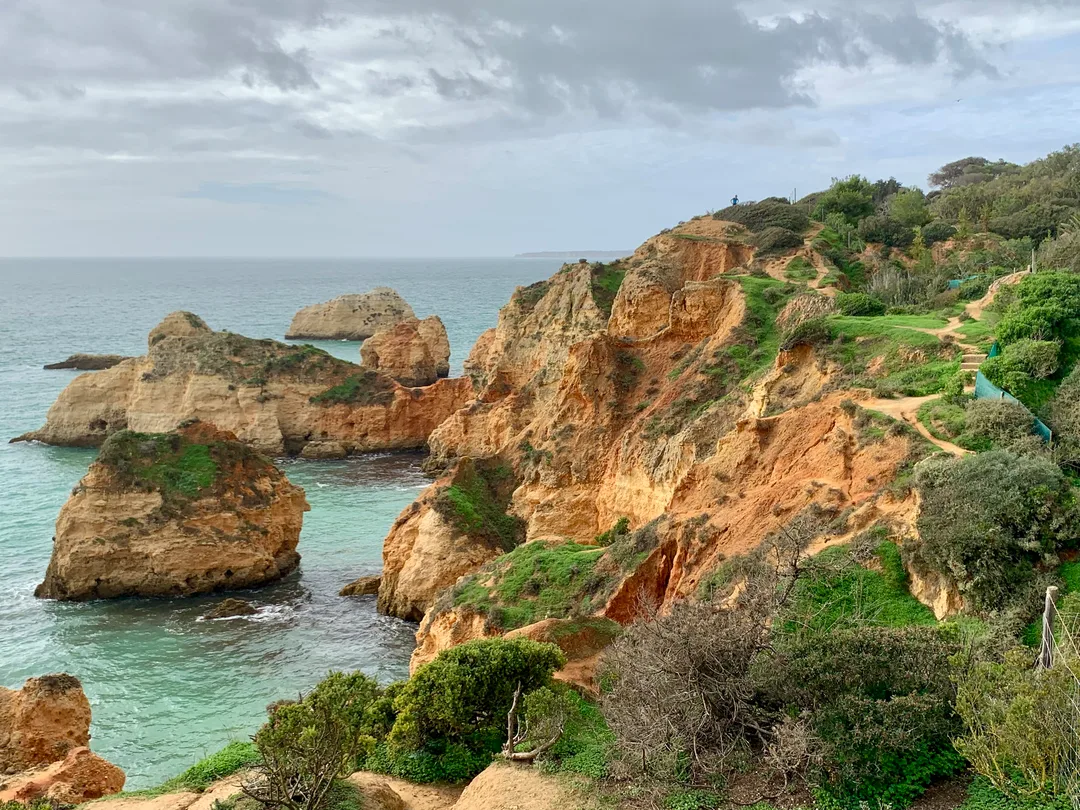 Sandy, orange colored bluffs with green vegetation overlooking blue water and grey sky.