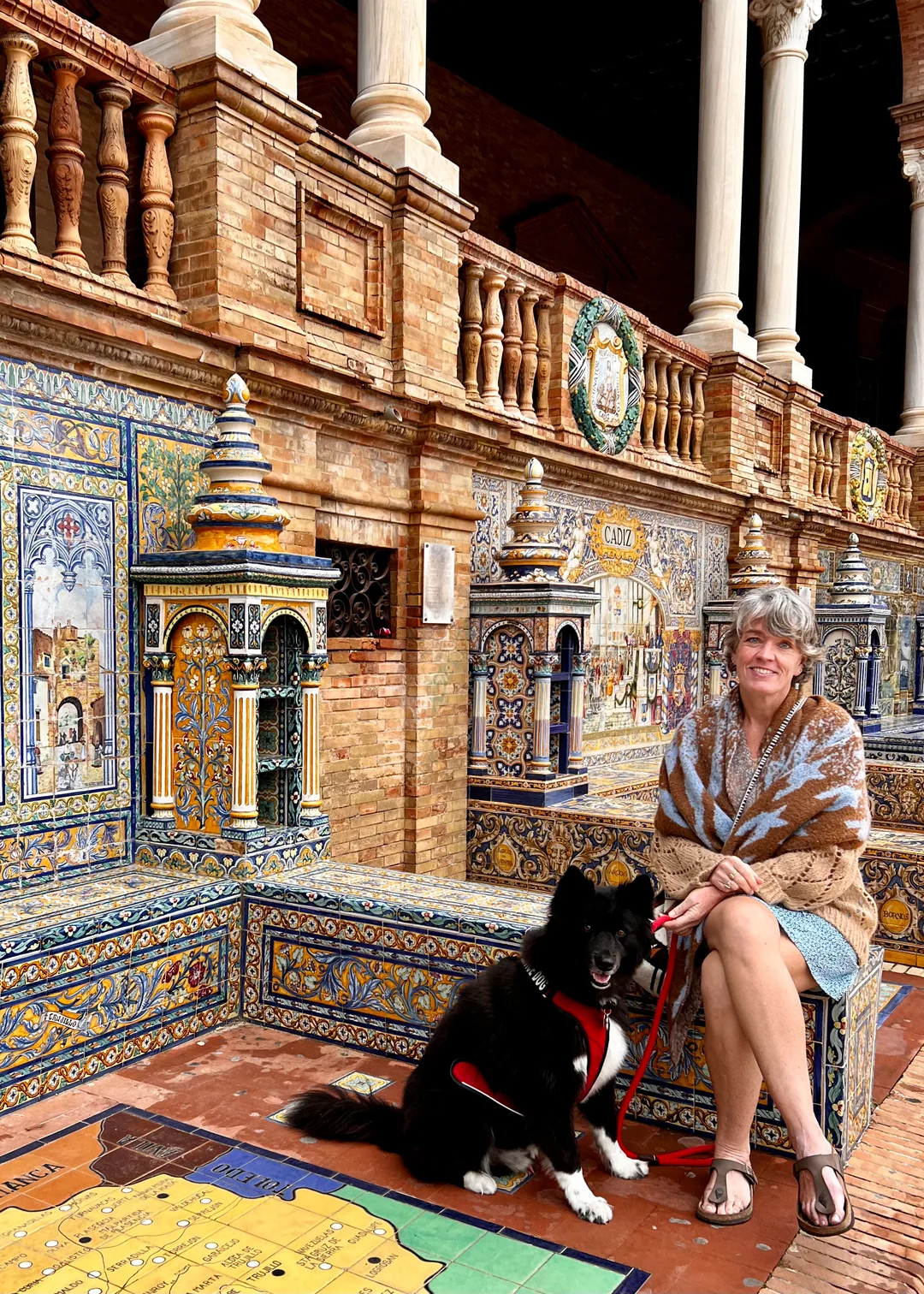 A woman and her dog sitting on a colorful tile bench next to a wall with a decorative tiled tower and colorful tiled scenes.