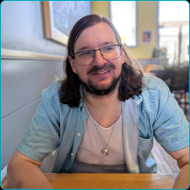 A man sitting at a table and facing the camera while his eyes are looking to the right. He has a slight grin. He's wearing a white tank top with a light blue unbuttoned shirt over it. He has a necklace with a silver ring hanging from it. His background is a cafe.